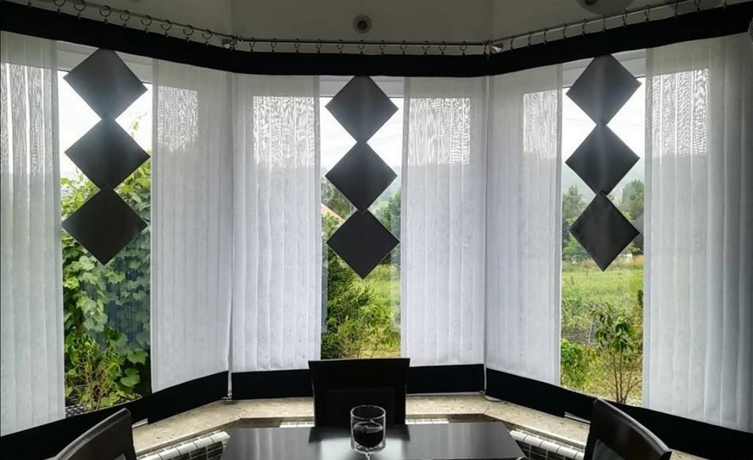 Nowoczesne, białe, pionowe żaluzje z czarnymi, geometrycznymi dekoracjami w kształcie rombów, widok na zielony ogród za oknem.