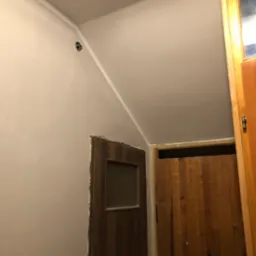 Świeżo wygładzona ściana w trakcie remontu, zabezpieczone gniazdka elektryczne żółtą taśmą malarską, widoczne dwie drewniane ościeżnice i fragment sufitu ze sztukaterią.