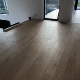 Ułożone panele podłogowe w jasnym odcieniu drewna w nowoczesnym wnętrzu z dużym oknem, widoczna puszka z farbą przy ścianie.