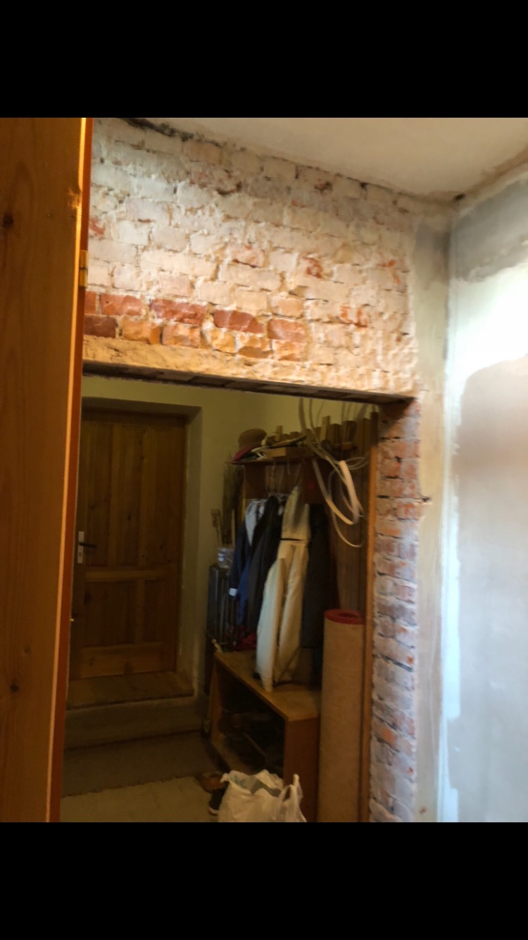 Częściowo otynkowana ściana z cegły w przejściu do przedpokoju z drewnianymi drzwiami i wieszakiem na ubrania, widoczne ślady prac remontowych.
