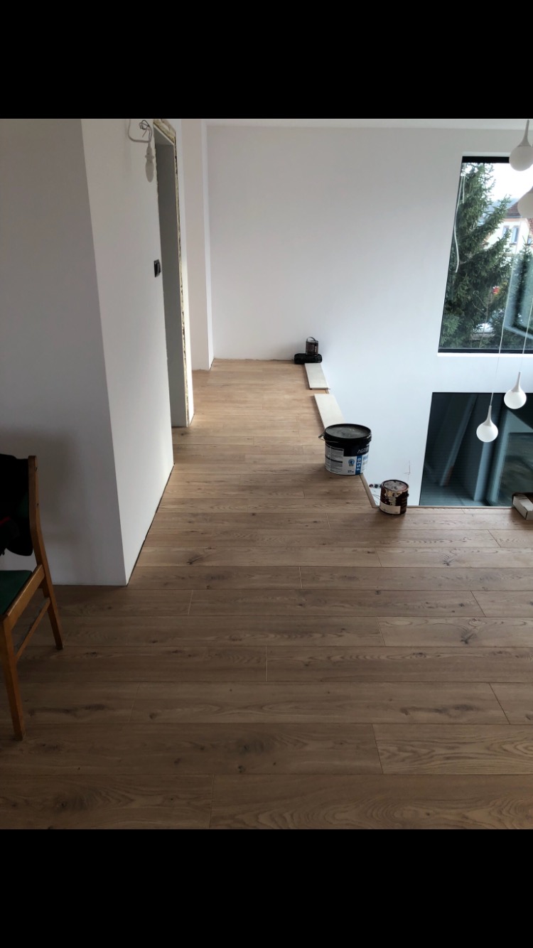 Ułożona drewniana podłoga w jasnym wnętrzu, widoczne puszki z farbą i fragment krzesła, wysokie okno z widokiem na drzewo na zewnątrz.