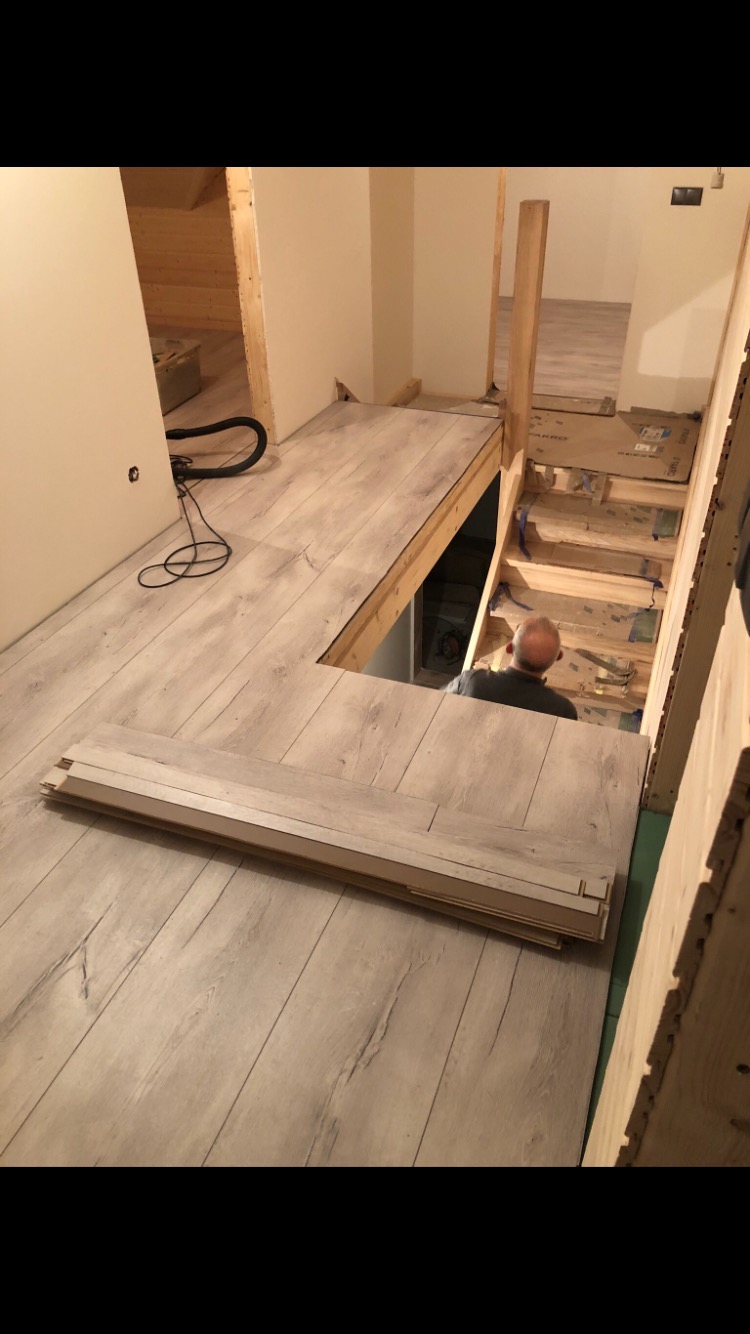 Fragment wnętrza w trakcie prac wykończeniowych: ułożona podłoga z paneli w odcieniach jasnego drewna, widoczne niedokończone schody z drewnianą konstrukcją oraz pracownik wchodzący na dół, paczka...