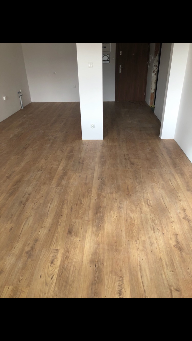 Nowo ułożone panele podłogowe w pustym pokoju z białymi ścianami i kolumną, widoczne gniazdka elektryczne. W tle drzwi wejściowe do mieszkania.