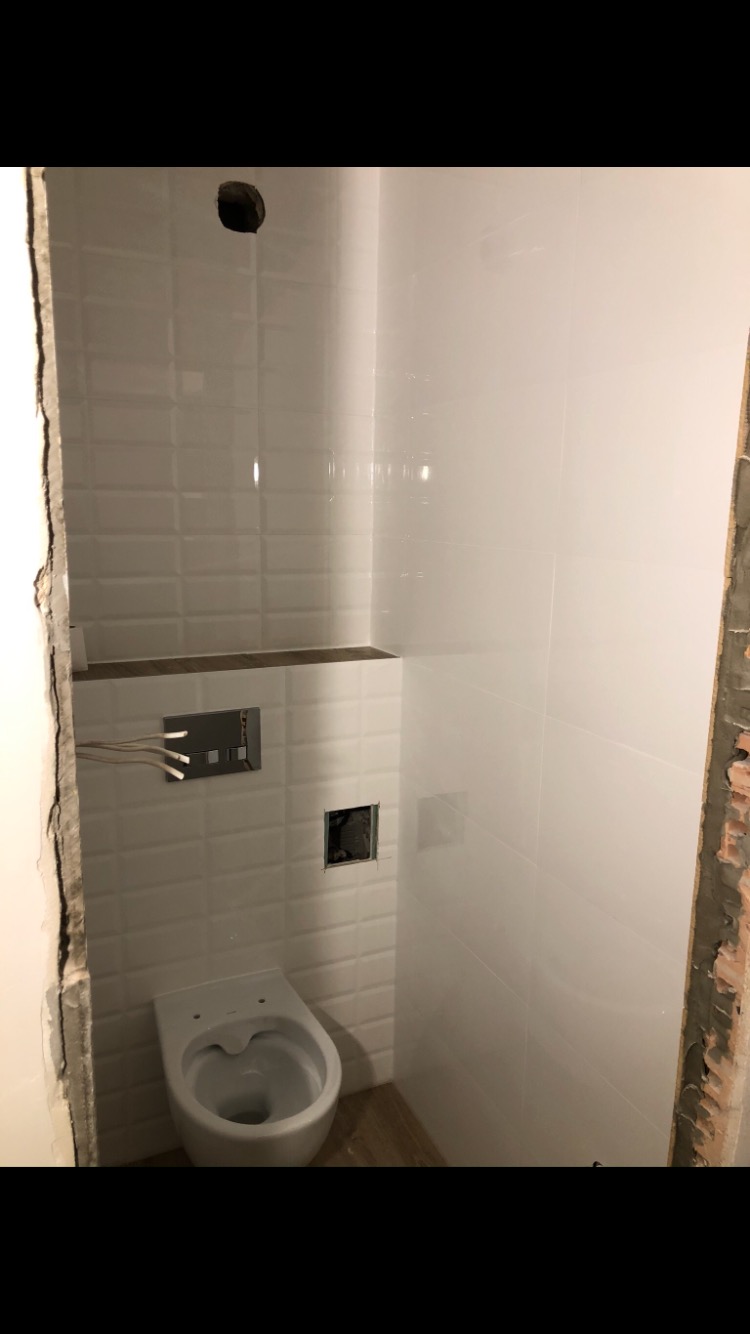 Wnętrze łazienki w trakcie remontu: białe płytki ułożone na ścianach, częściowo zamontowana toaleta podwieszana, widoczne instalacje wodne i elektryczne, surowe wykończenie ściany po lewej stronie.