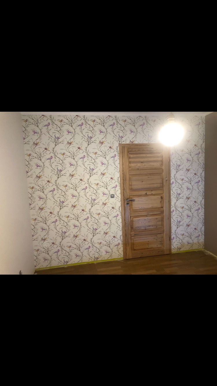 Pokój po remoncie: ściana wykończona tapetą w ptaki i gałęzie, drewniane drzwi z poziomymi panelami, widoczne zabezpieczenia podłogi taśmą malarską.