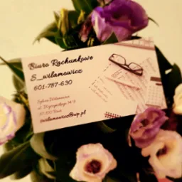 Wizytówka biura rachunkowego Sylwia Wilamowicz umieszczona w bukiecie z fioletowo-białych kwiatów, obok kalkulator, okulary i dokumenty z tabelami.