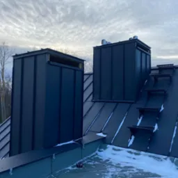 Nowoczesny, czarny dach z blachy na rąbek stojący, z dwoma kominami obłożonymi blachą i stopniami dachowymi, częściowo pokryty śniegiem.