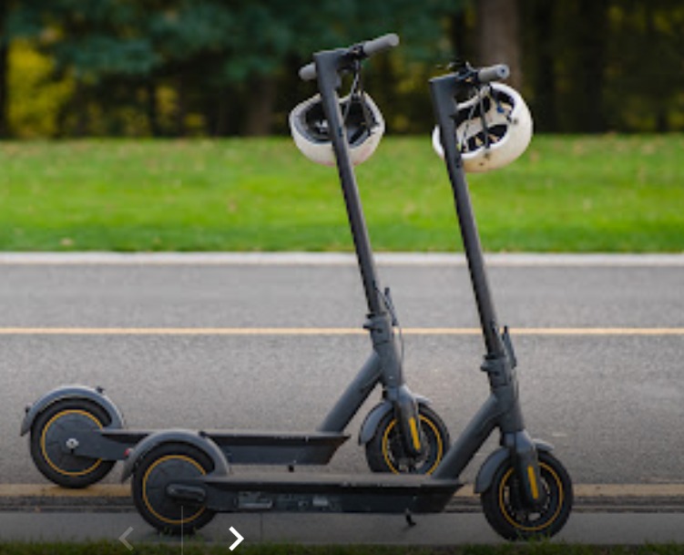 Dwa elektryczne skutery stojące obok siebie na asfalcie, z białymi kaskami zawieszonymi na kierownicach, tło z zielenią.