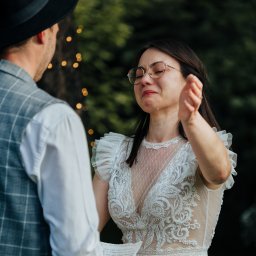 KOMP-BOX Urszula Gajewska - Panna młoda w okularach, ze łzami w oczach, w koronkowej sukni ślubnej podczas ceremonii plenerowej. W tle zieleń i dekoracyjne lampki.