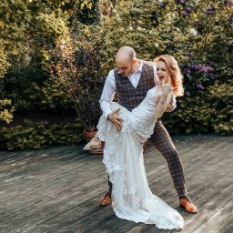 KOMP-BOX Urszula Gajewska - Uśmiechnięta panna młoda w koronkowej sukni niesiona przez pana młodego w kraciastej kamizelce na tle zieleni i drewnianego tarasu.