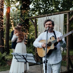 KOMP-BOX Urszula Gajewska - Muzyk z gitarą śpiewa na ślubie na tle pary młodej w objęciach, oświetlonej girlandami. Scena z drewnianą konstrukcją i makramą w tle, nuty na statywie.