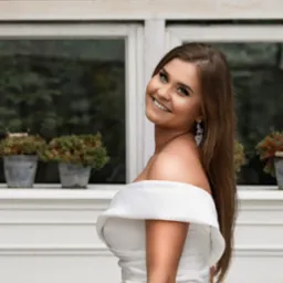 Uśmiechnięta brunetka w białej sukni z odkrytymi ramionami, pozuje na tle białego okna z doniczkami. Eleganckie kolczyki.