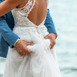 Para młoda: mężczyzna w błękitnej marynarce obejmuje kobietę w koronkowej sukni ślubnej z odkrytymi plecami na tle wody. Widoczne detale sukni i biżuteria.