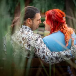 Para zakochanych, obejmująca się, patrzy sobie w oczy. Mężczyzna w kwiecistej koszuli, kobieta z płomiennymi, rudymi włosami, upiętymi ozdobną spinką.