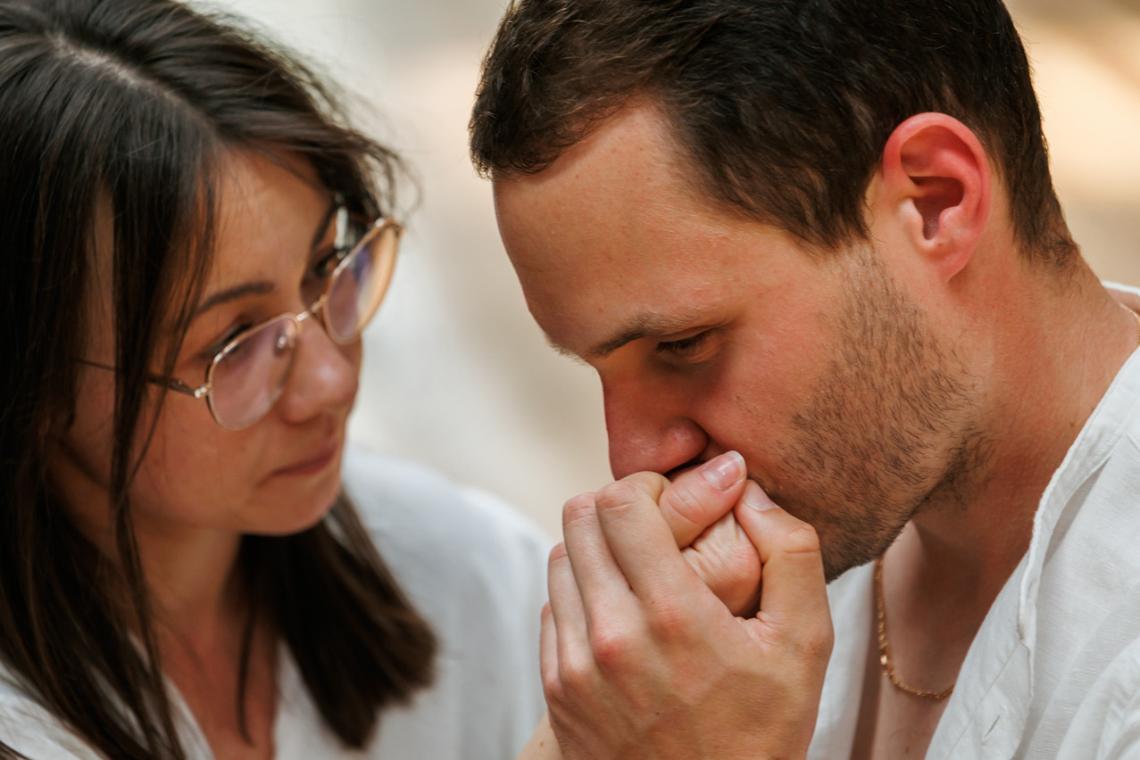 Delikatne ujęcie: mężczyzna całuje dłoń kobiety w okularach, oboje w białych koszulach, subtelne oświetlenie, złoty łańcuszek mężczyzny dodaje blasku.