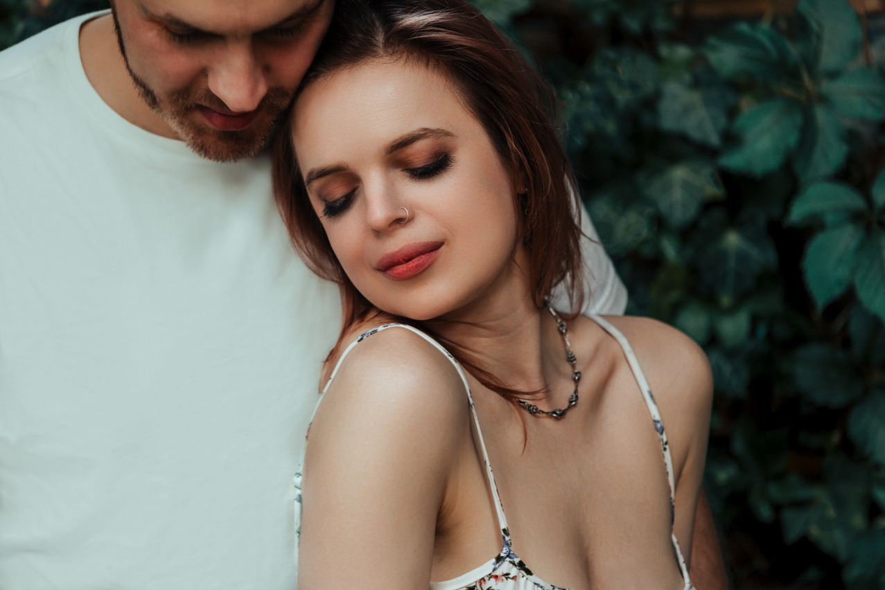 Romantyczna sesja narzeczeńska: zakochana para w objęciach na tle zieleni, ona w kwiecistej sukience z delikatną biżuterią, on w białym t-shircie.