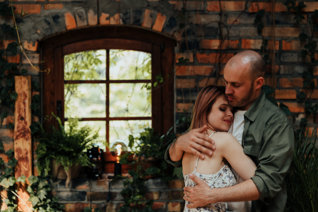 Zakochana para obejmuje się na tle ceglanej ściany z oknem i zielenią. Mężczyzna w zielonej koszuli, kobieta w kwiecistej sukience. Ciepłe oświetlenie, romantyczny nastrój.