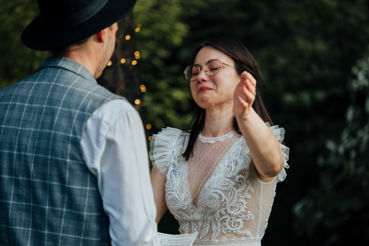 Panna młoda w okularach, ze łzami w oczach, w koronkowej sukni ślubnej podczas ceremonii plenerowej. W tle zieleń i dekoracyjne lampki.