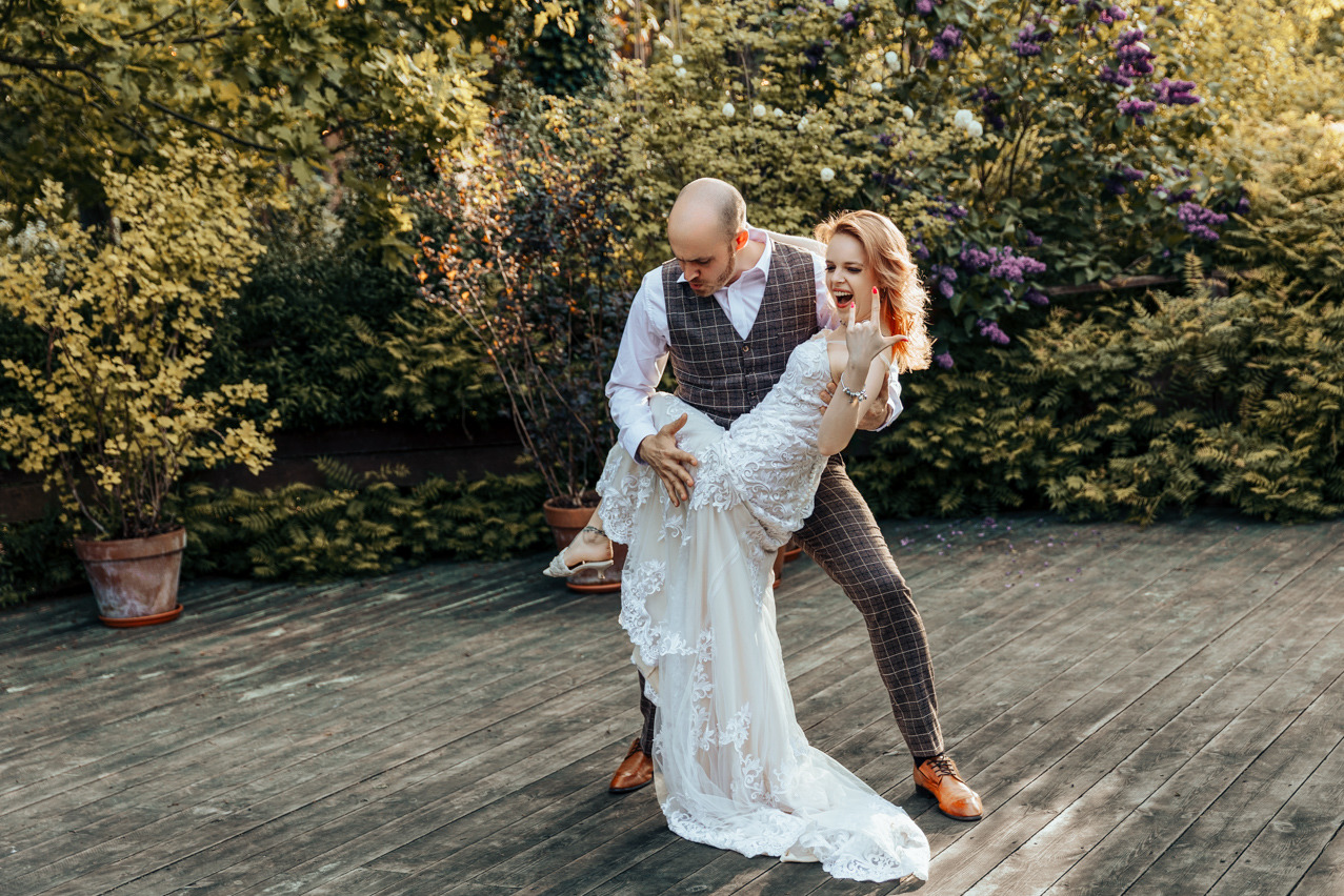 Uśmiechnięta panna młoda w koronkowej sukni niesiona przez pana młodego w kraciastej kamizelce na tle zieleni i drewnianego tarasu.