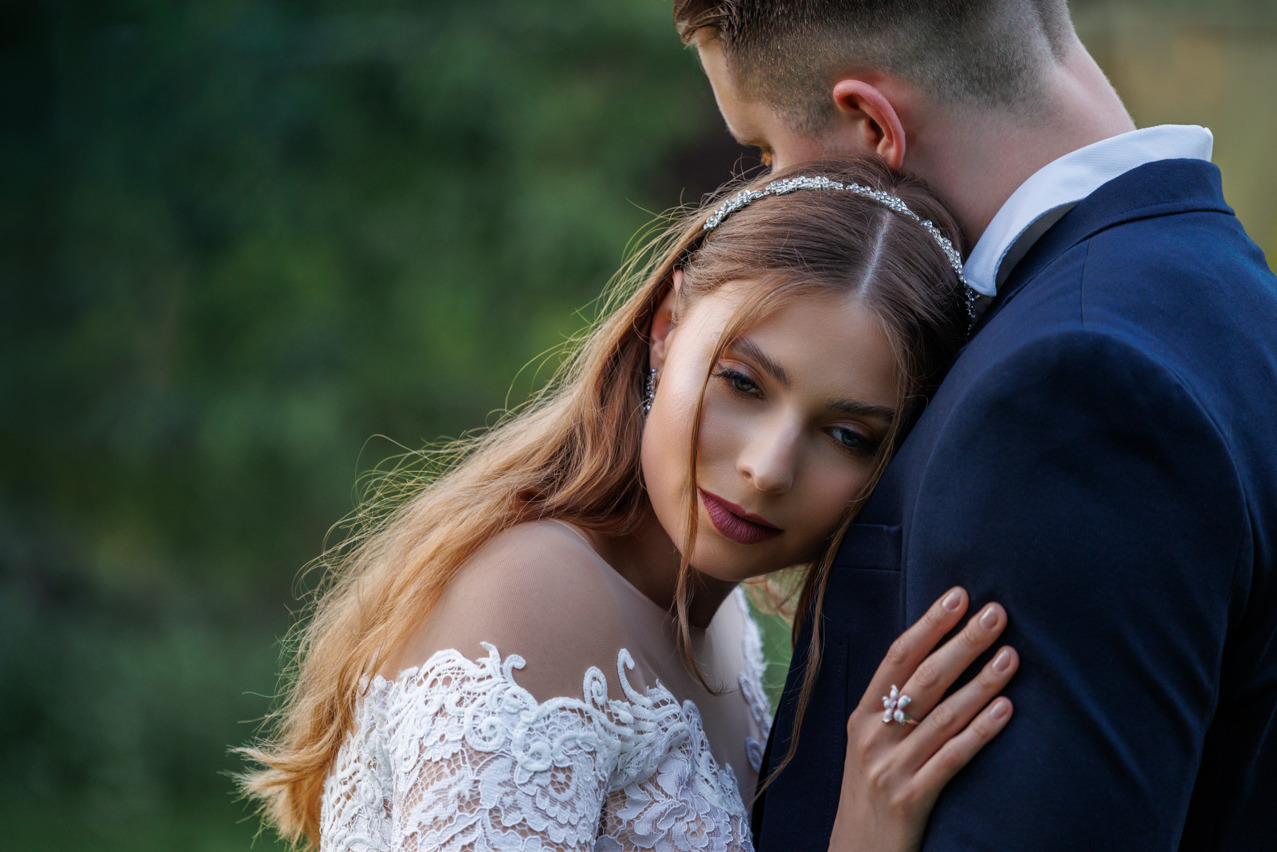 Romantyczne ujęcie panny młodej w koronkowej sukni, opierającej głowę o ramię pana młodego. Delikatna biżuteria i stonowany makijaż podkreślają subtelność chwili.