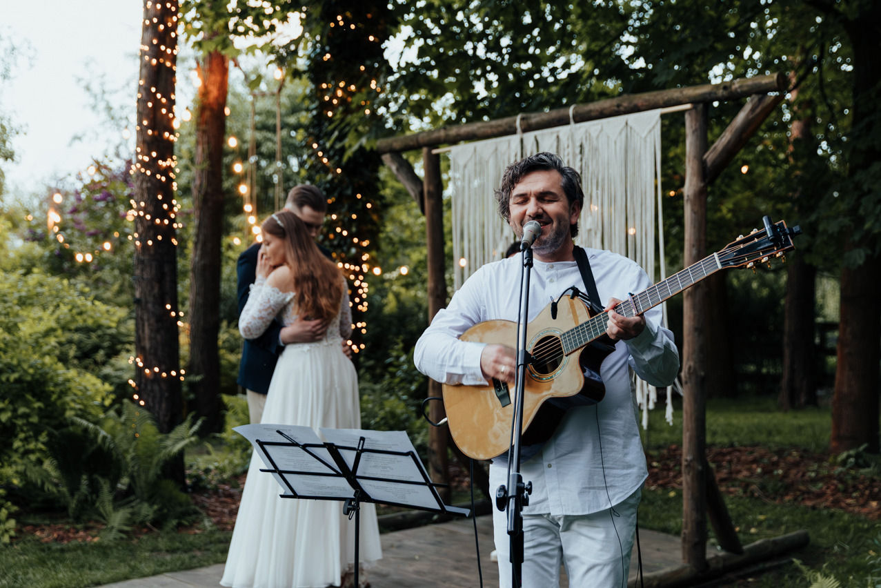 Muzyk z gitarą śpiewa na ślubie na tle pary młodej w objęciach, oświetlonej girlandami. Scena z drewnianą konstrukcją i makramą w tle, nuty na statywie.