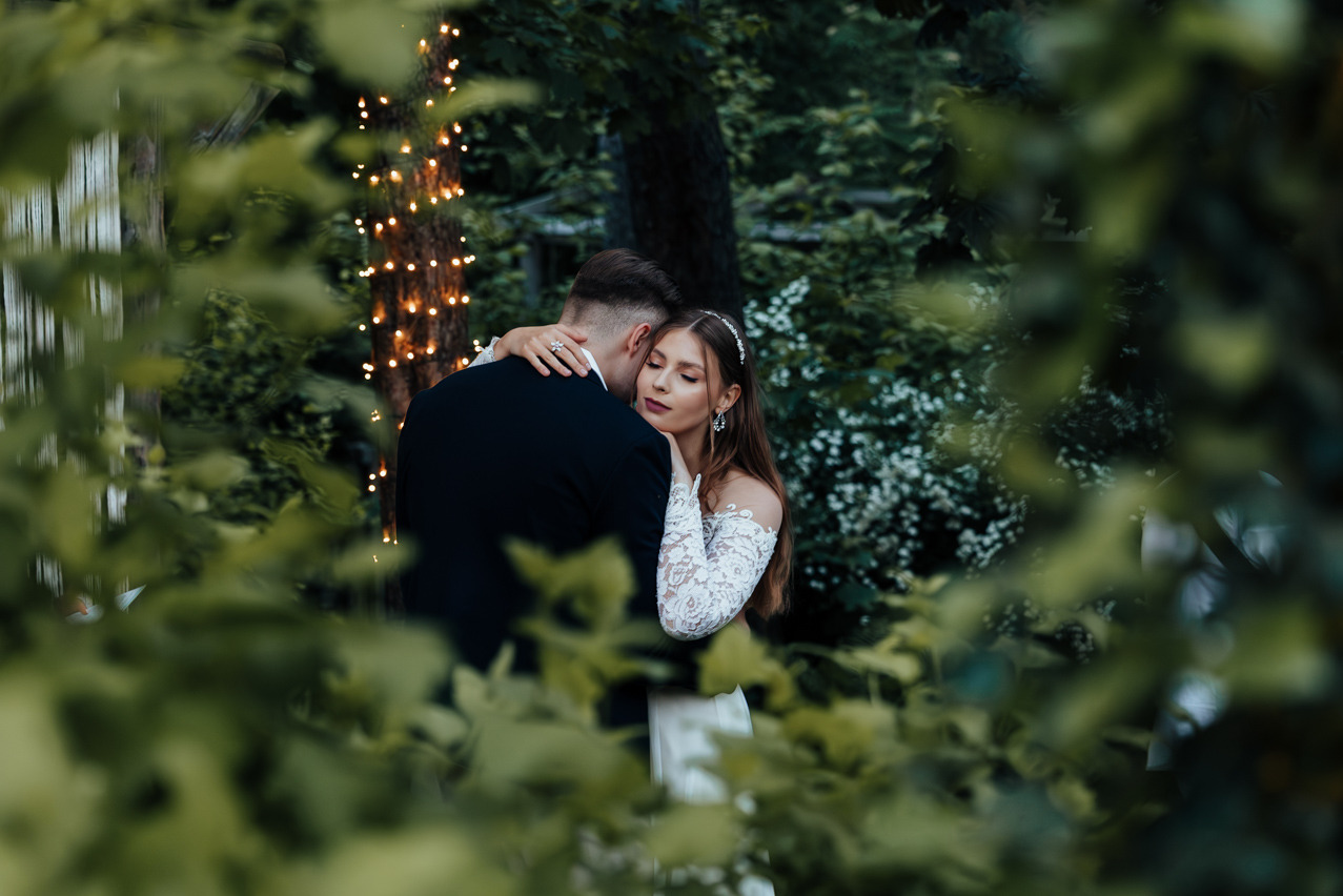 Para młoda w objęciach, on w czarnym garniturze, ona w białej sukni z koronkowymi rękawami, na tle drzewa owiniętego lampkami, ujęcie przez liście.