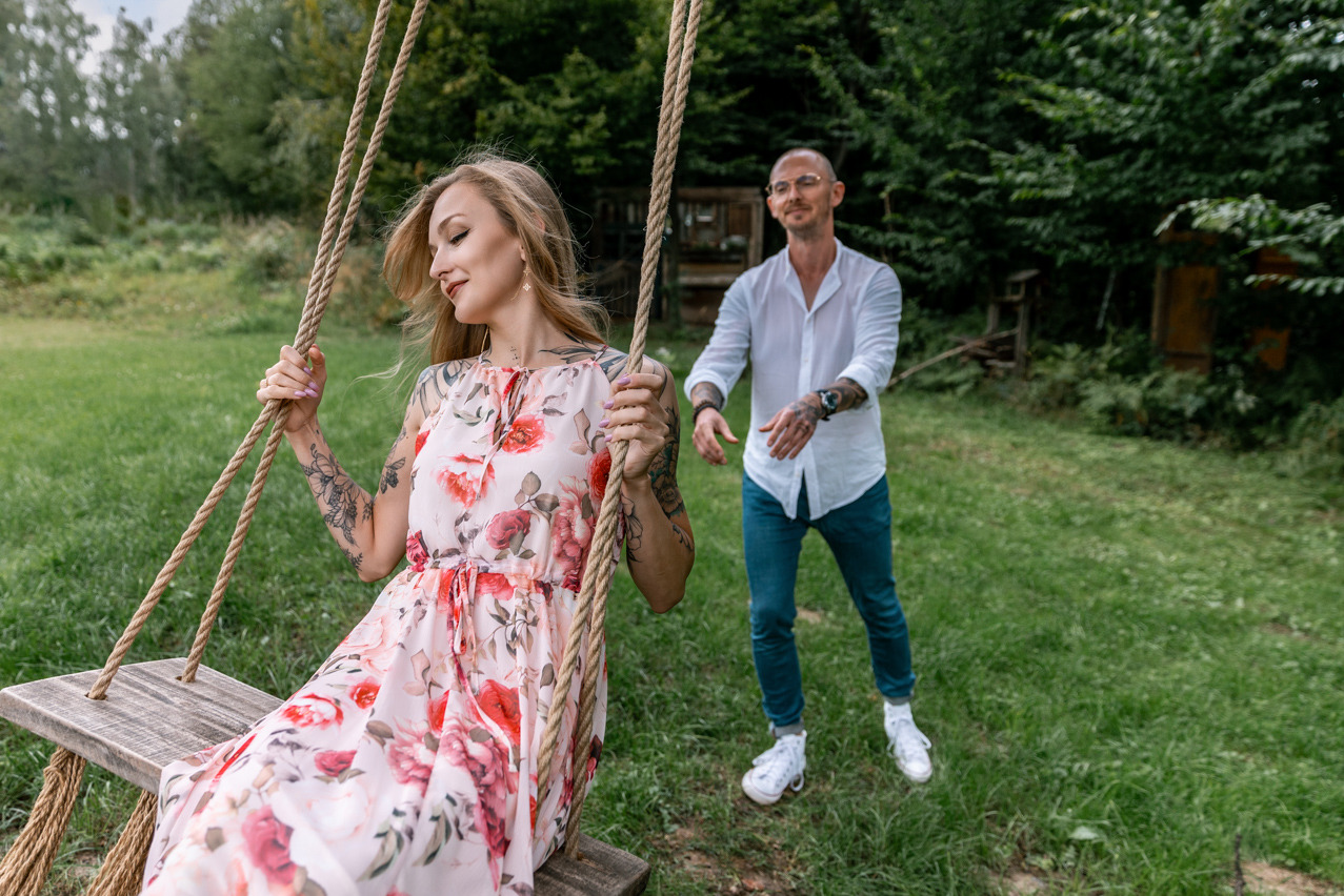 Para zakochanych na huśtawce: kobieta w kwiecistej sukience i mężczyzna w białej koszuli, oboje z tatuażami, w scenerii zielonego ogrodu. Romantyczna atmosfera.