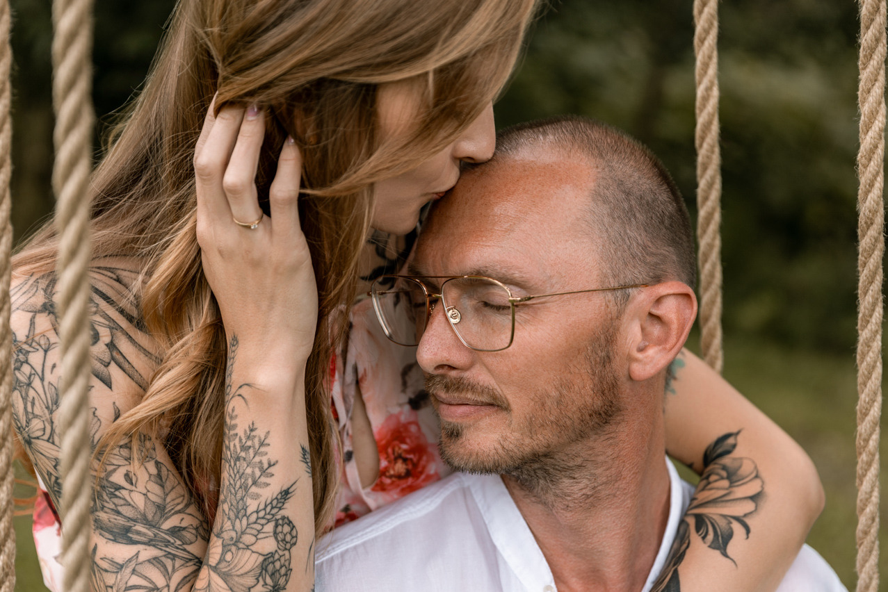 Romantyczne ujęcie pary na huśtawce: kobieta z tatuażami na ramionach całuje mężczyznę w okularach w czoło, zieleń w tle.