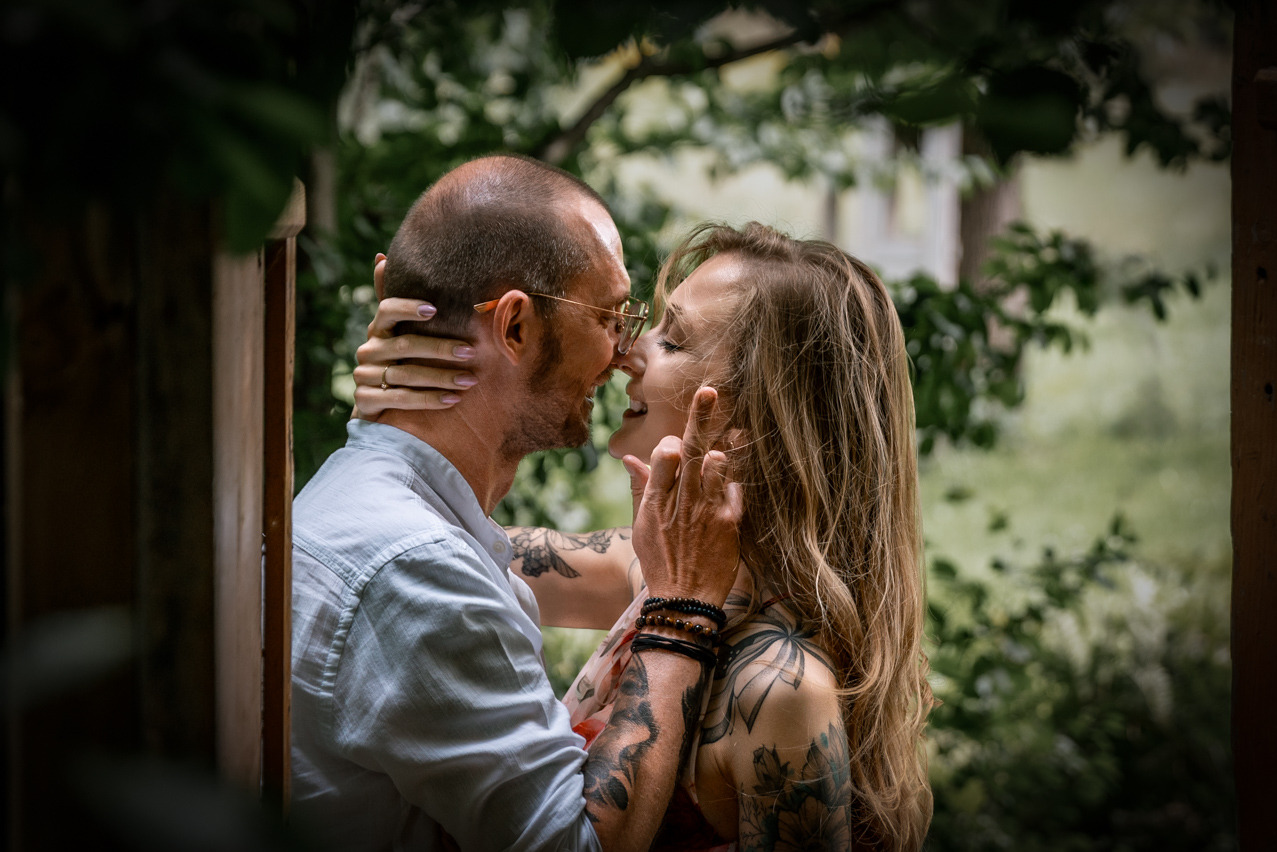 Zakochana para, on w okularach i koszuli, ona z długimi włosami i tatuażami, czule dotykają się nosami na tle zieleni, uchwyceni w romantycznym ujęciu.