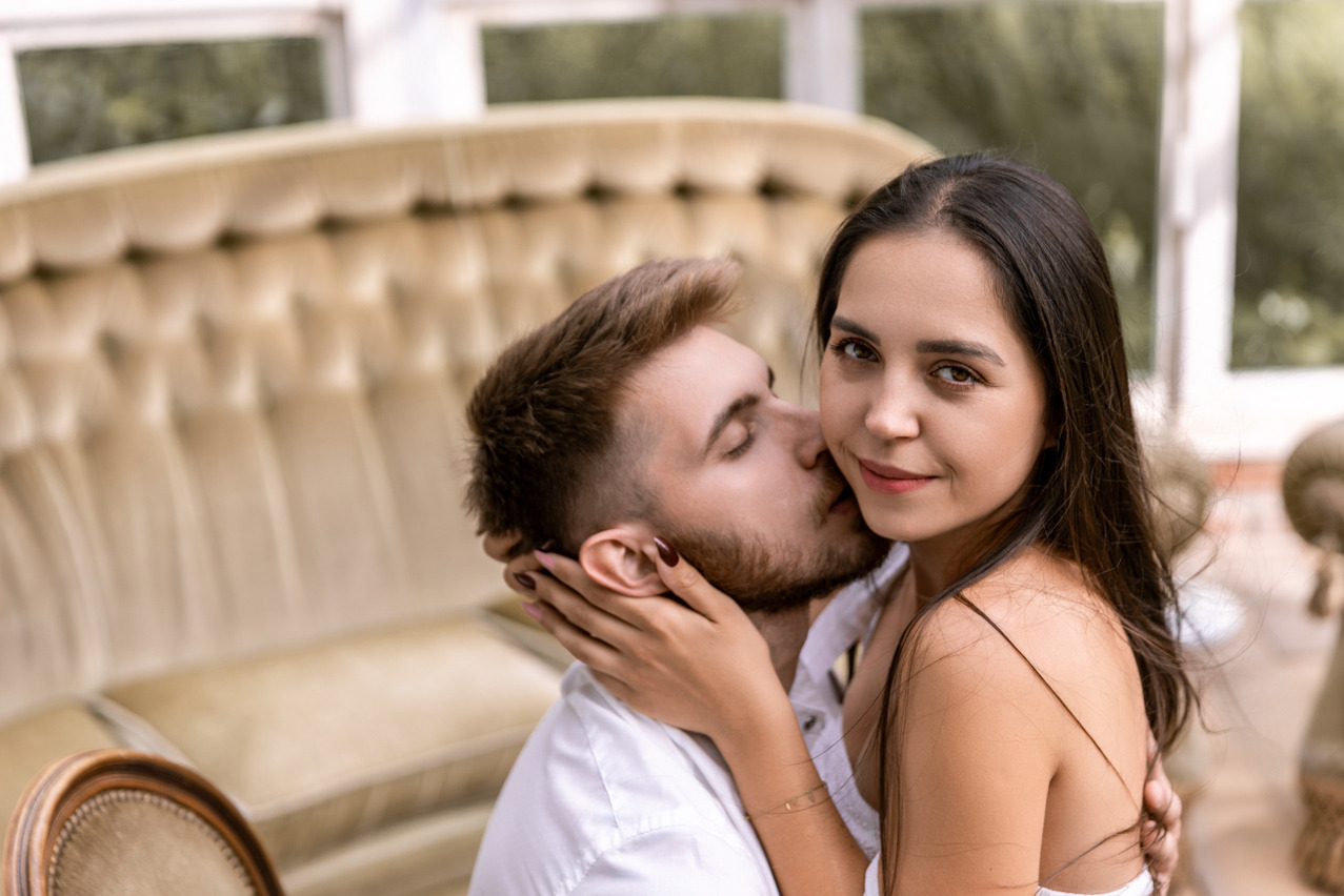 Zakochana para: mężczyzna całuje kobietę w szyję, ona spogląda w obiektyw z uśmiechem. W tle kremowa sofa i jasne wnętrze. Romantyczna sesja w plenerze.