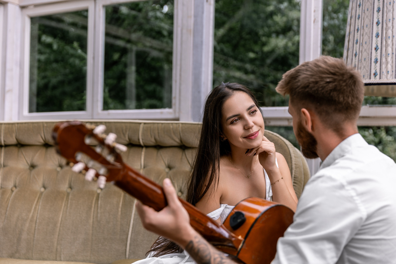 Uśmiechnięta brunetka w białej sukience słucha gitarzysty w jasnym wnętrzu z oknami i zielenią w tle, w tle widoczna lampa.