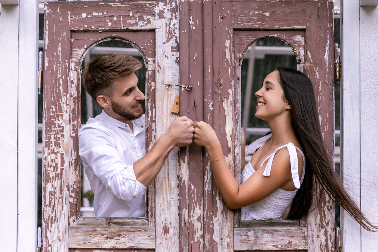 Uśmiechnięta para w białych strojach, złączone pięści przez stare, rozpadające się drewniane drzwi z łuszczącą się farbą, romantyczna sesja w plenerze.