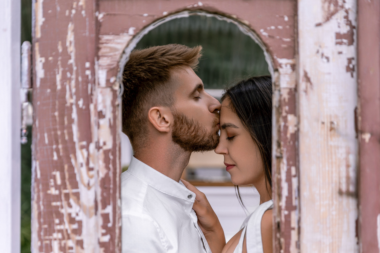 Mężczyzna w białej koszuli całuje kobietę w czoło, oboje zamknięte oczy, ujęcie przez starą, łuszczącą się ramę okienną.