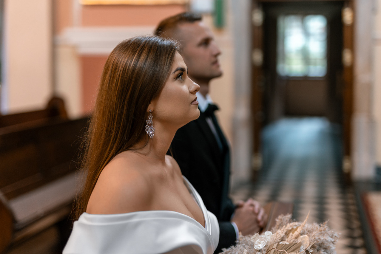 Para młoda w kościele: panna młoda z błyszczącymi kolczykami, pan młody w tle. Ujęcie z profilu, skupione na szczegółach stroju i dekoracji.