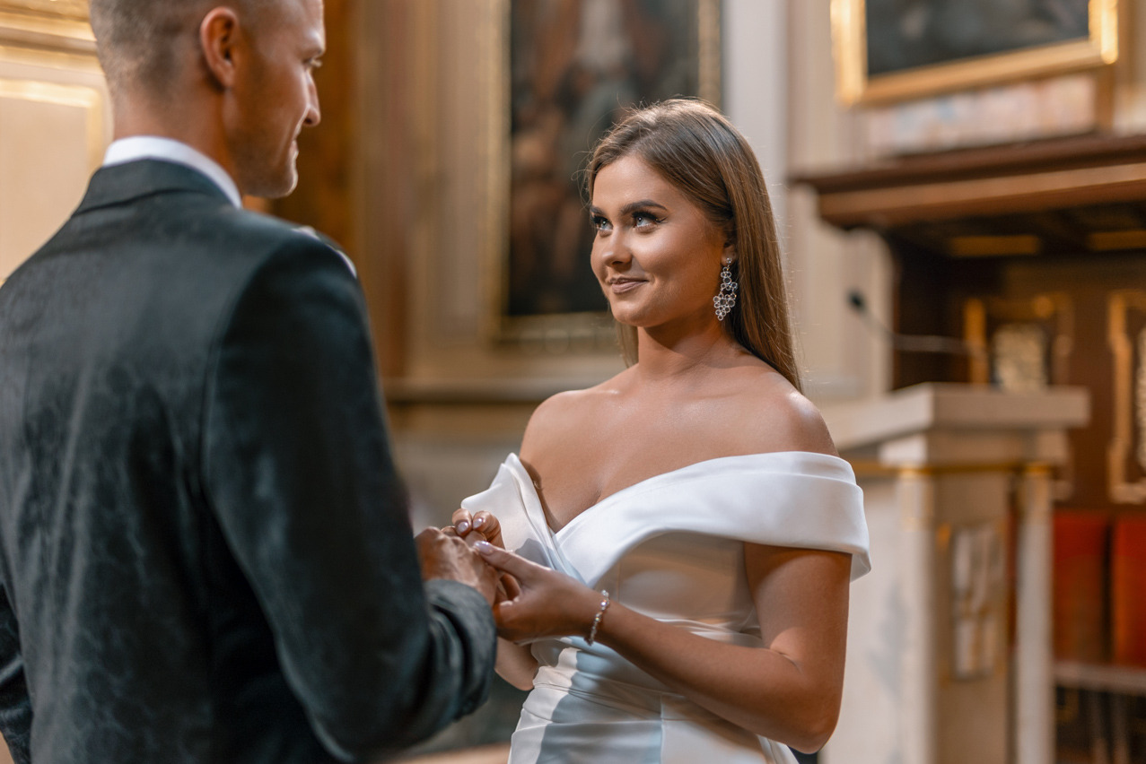Panna młoda w białej sukni, z błyszczącymi kolczykami, uśmiecha się, patrząc na pana młodego podczas ceremonii ślubnej w eleganckim wnętrzu kościoła.