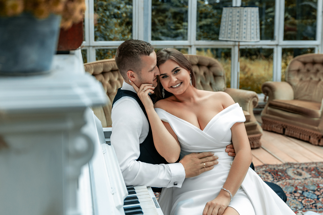 Uśmiechnięta para narzeczeńska w eleganckim wnętrzu z białym pianinem. Ona w białej sukni, on w kamizelce. Romantyczna sesja w jasnym, przestronnym pomieszczeniu.