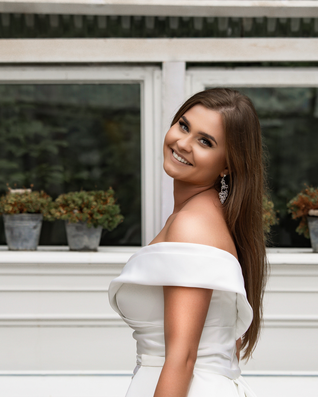 Uśmiechnięta brunetka w białej sukni z odkrytymi ramionami, pozuje na tle białego okna z doniczkami. Eleganckie kolczyki.