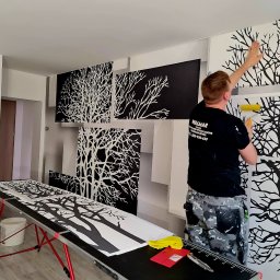 WALMAR uslugi budowlano wykonczeniowe Waldemar Nowak - Pracownik w czarnej koszulce z logo firmy nakłada wałkiem tapetę z motywem drzewa na ścianę, widoczne panele tapety ułożone na stole, narzędzia pracy i wiadro z klejem.