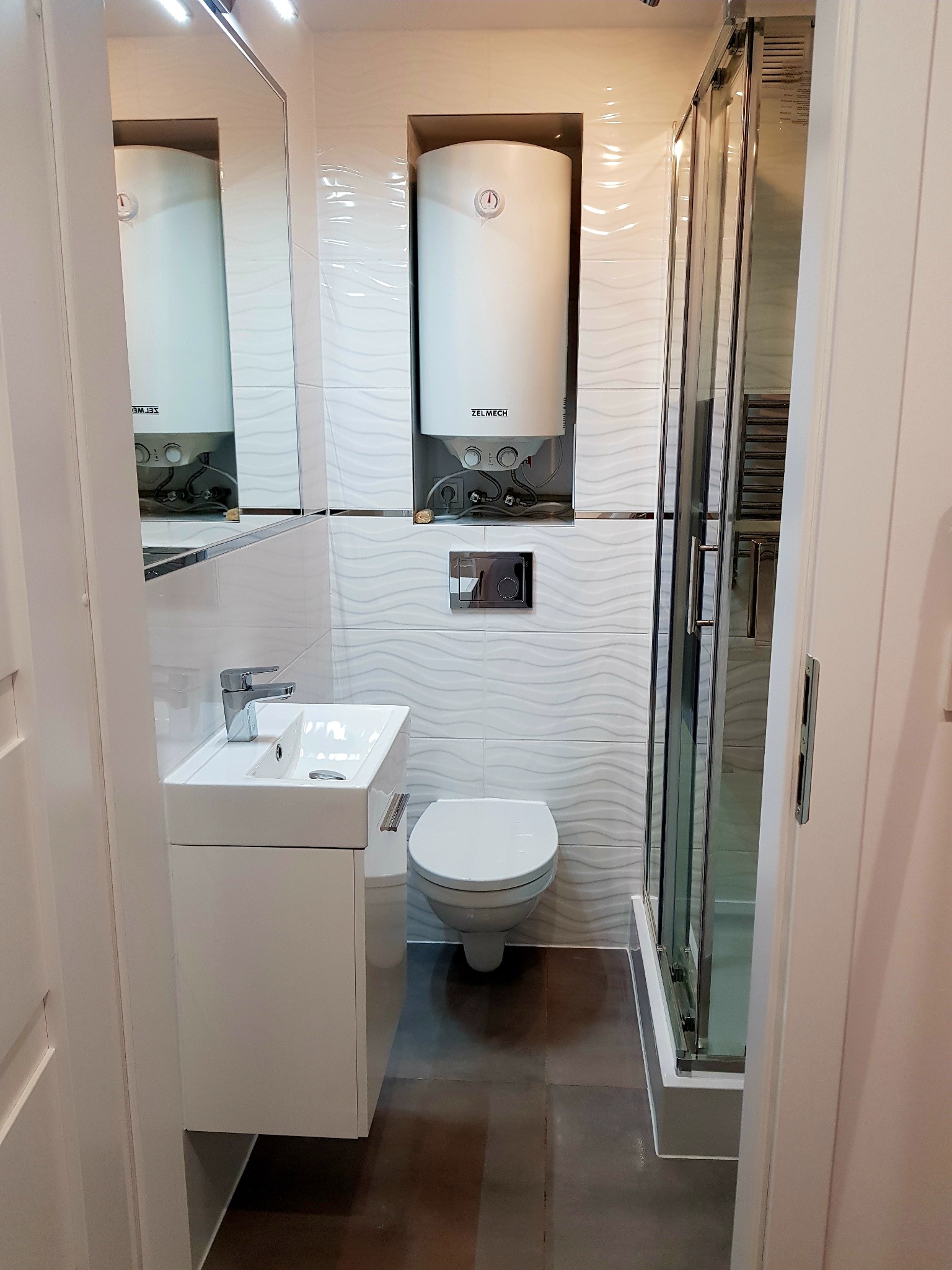 Nowoczesna łazienka z białymi płytkami o falistej strukturze, wiszącą szafką z umywalką, podgrzewaczem wody i kabiną prysznicową z przesuwnymi drzwiami, widok z wejścia.