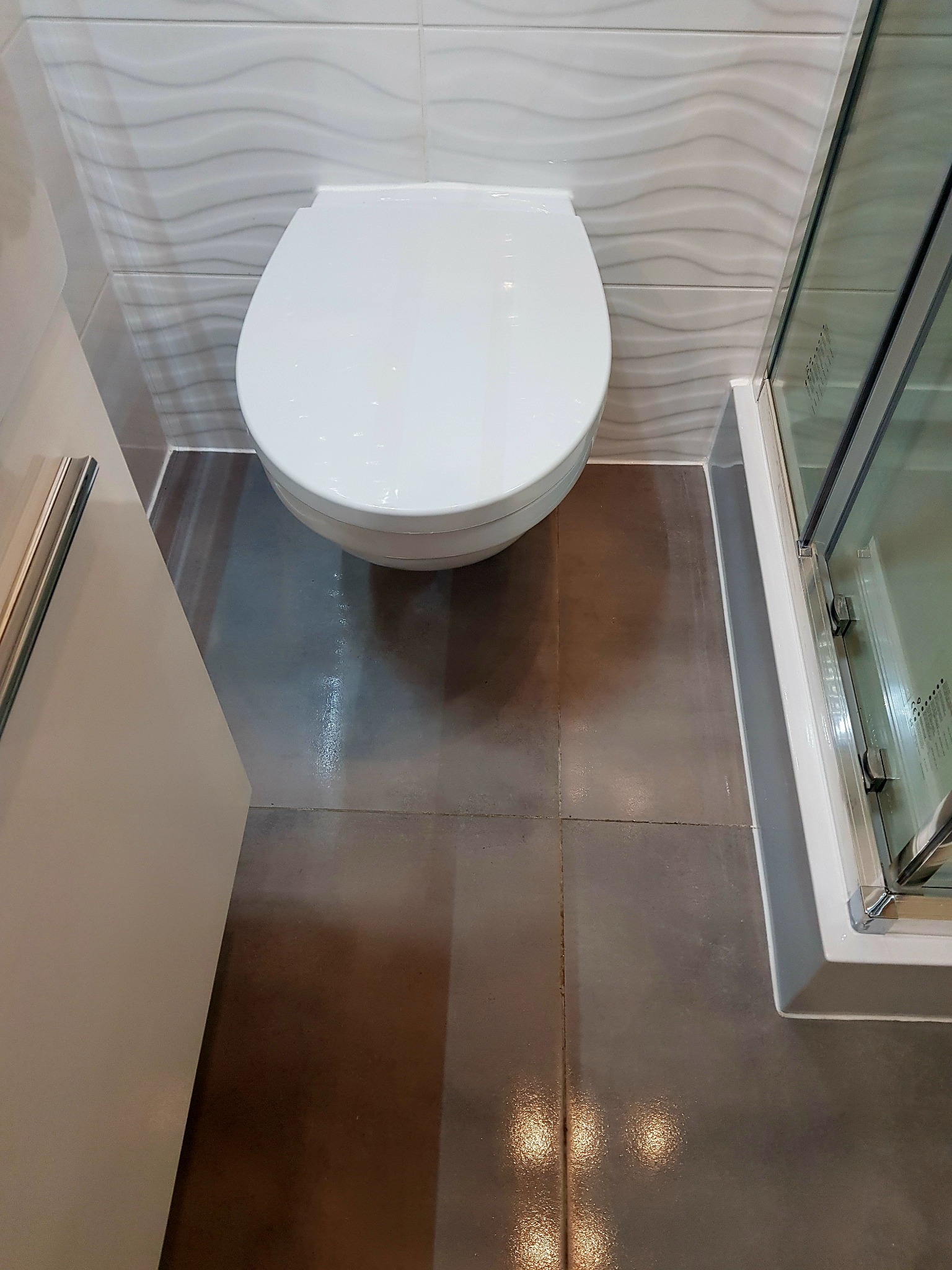 Nowoczesna łazienka po remoncie: podwieszana toaleta, szare płytki podłogowe, białe płytki ścienne z delikatnym wzorem fal, kabina prysznicowa z chromowanymi zawiasami.