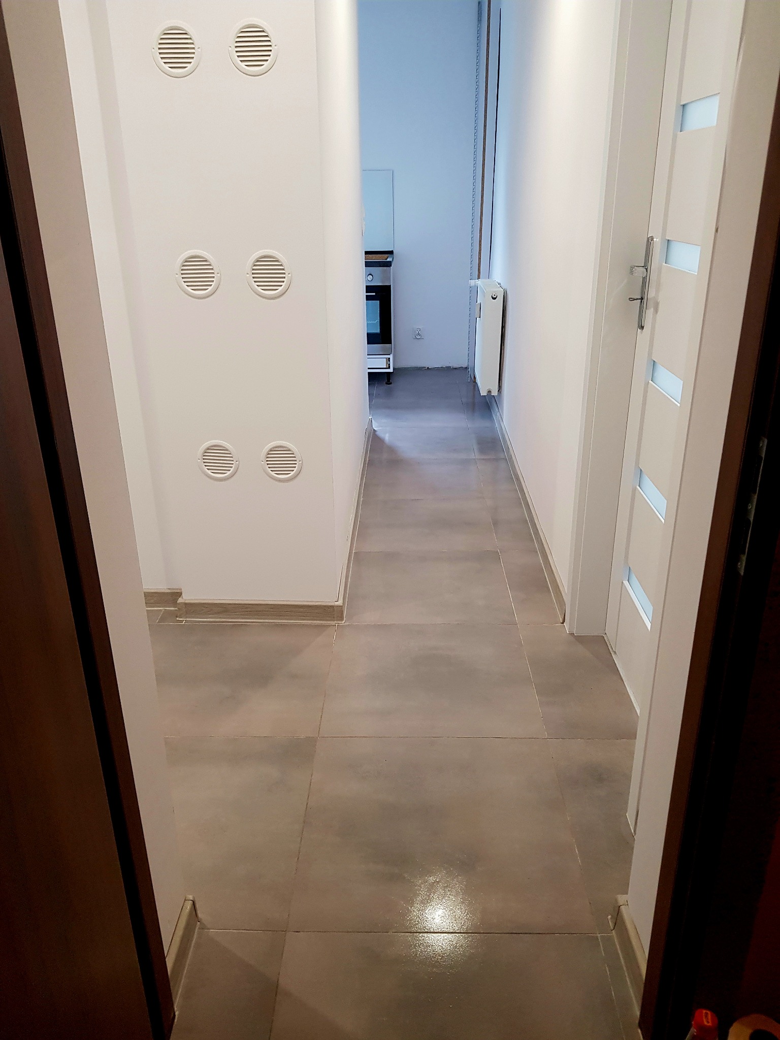 Wykończony korytarz z szarą podłogą z płytek, białe ściany z czterema okrągłymi kratkami wentylacyjnymi i widokiem na kuchnię w tle.