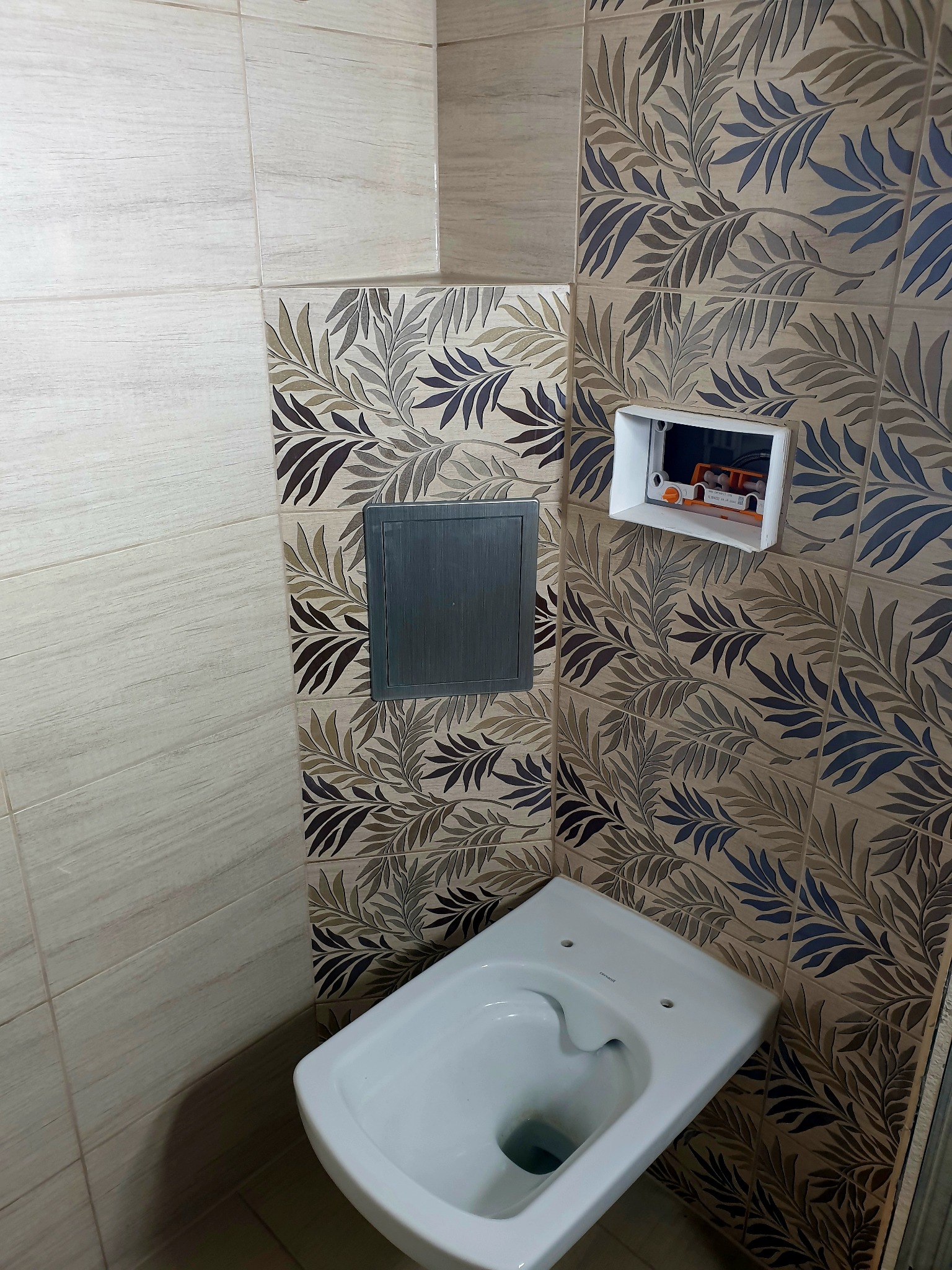 Wisząca miska WC zamontowana na tle płytek z motywem liści, widoczna skrzynka instalacyjna i rewizja kanalizacyjna.