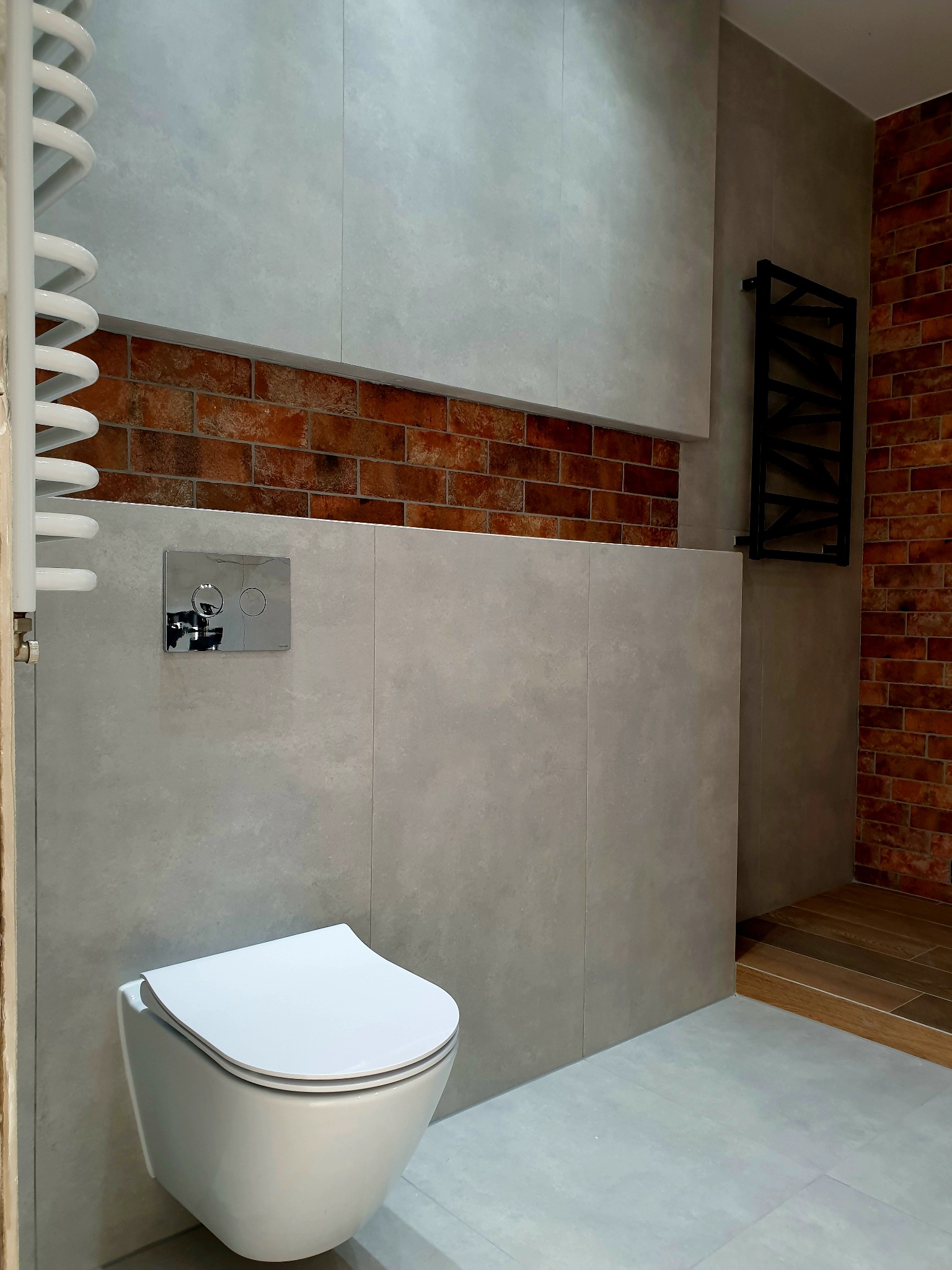 Nowoczesna łazienka z wiszącą toaletą, szarymi płytkami imitującymi beton, akcentem z cegły i czarnym grzejnikiem drabinkowym.