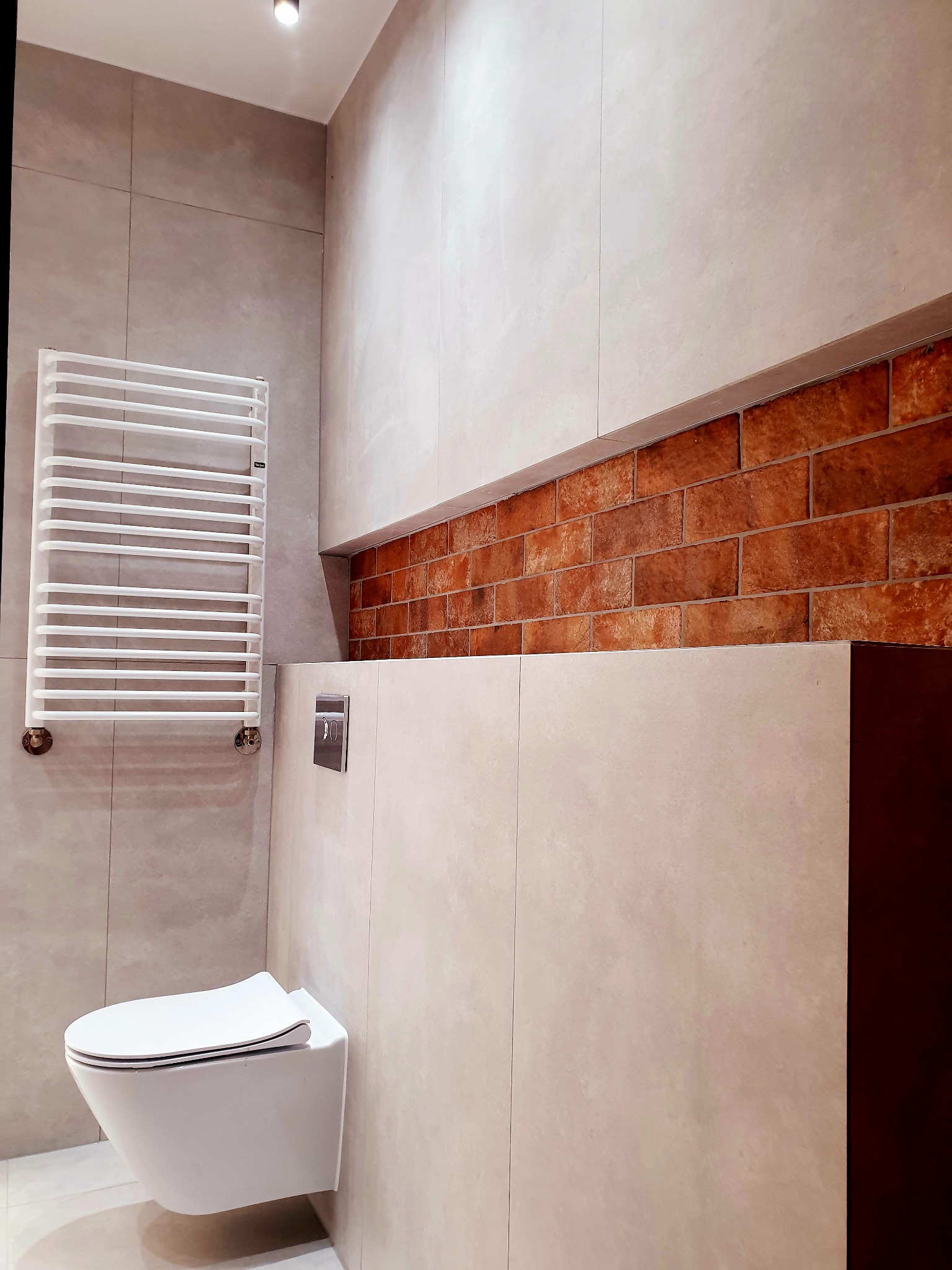Nowoczesna łazienka z podwieszaną toaletą, grzejnikiem drabinkowym i akcentem z cegły nad zabudową stelaża.