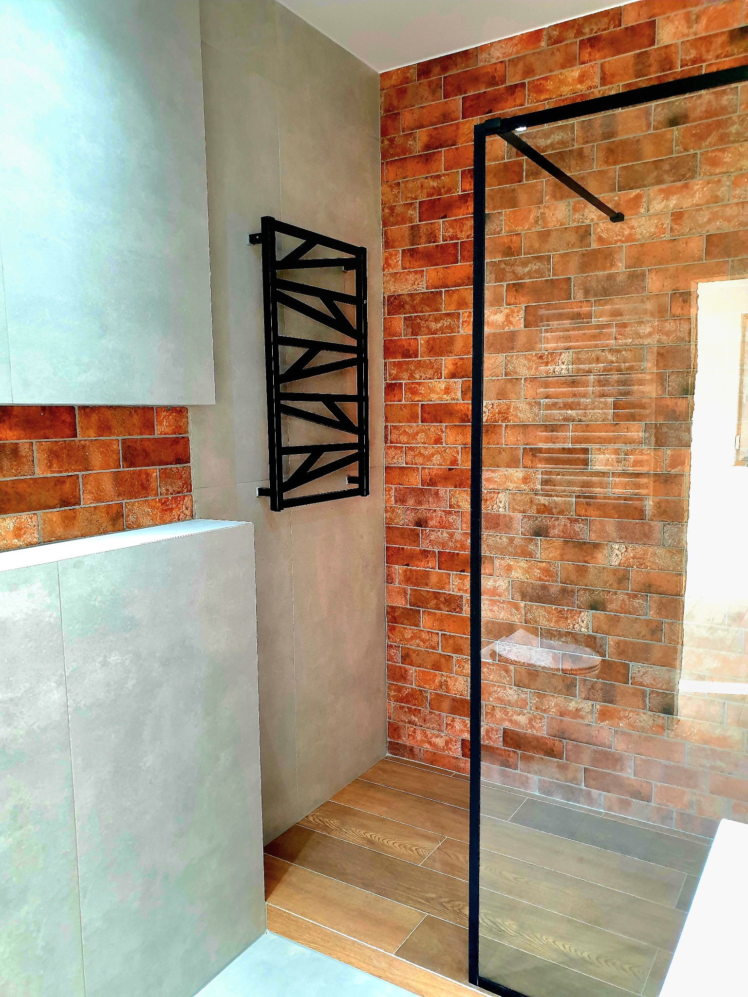 Nowoczesna łazienka z kabiną prysznicową typu walk-in, ściana wyłożona cegiełką w odcieniach czerwieni i brązu, kontrastująca z szarą płytą, drewnopodobna podłoga, czarny grzejnik drabinkowy.