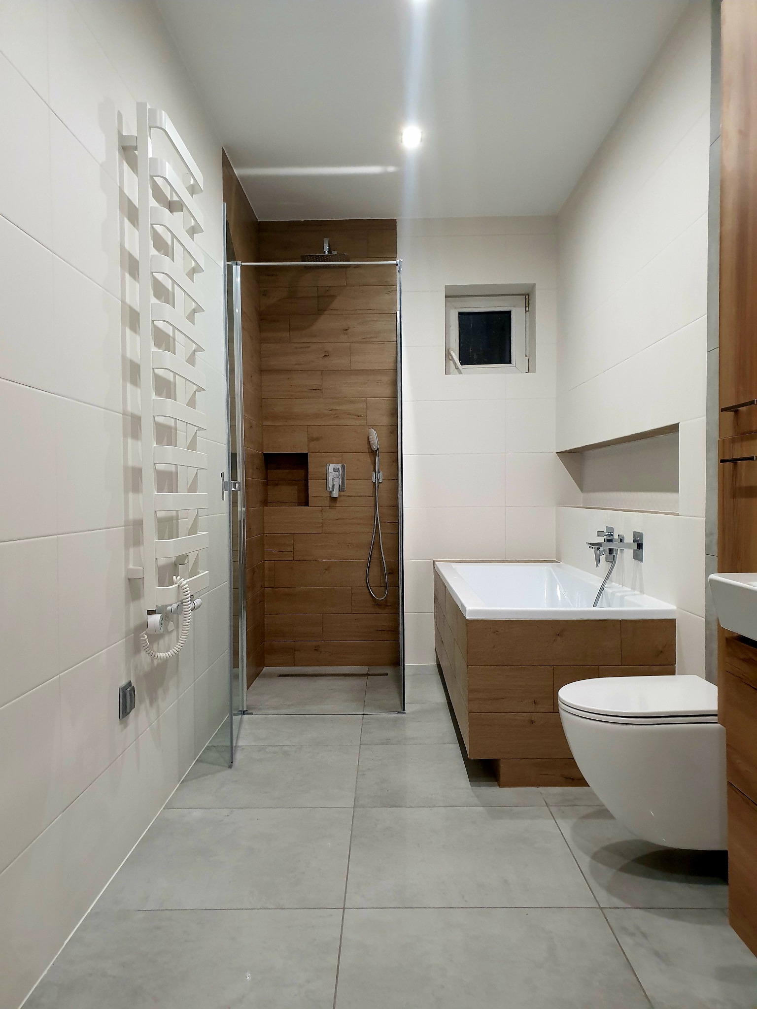 Nowoczesna łazienka z kabiną prysznicową wyłożoną drewnopodobnymi płytkami, białą wanną w obudowie z drewna i wiszącą toaletą.