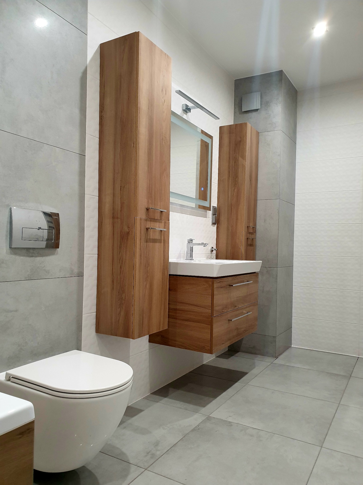 Nowoczesna łazienka z wiszącą toaletą, drewnianą szafką pod umywalką i dwoma wysokimi szafkami, szare płytki na ścianach i podłodze, oświetlone lustro nad umywalką, chromowana armatura.
