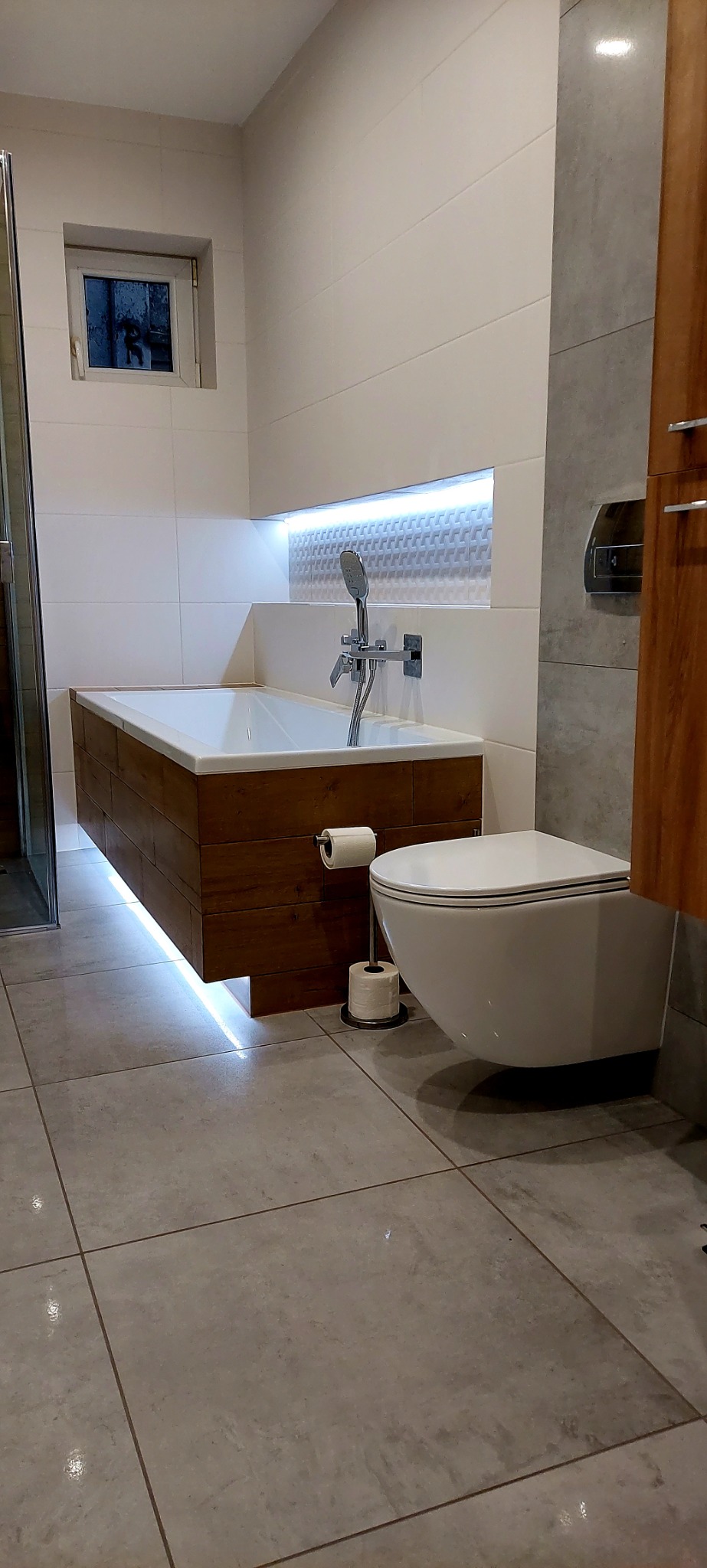 Nowoczesna łazienka z podświetlaną wanną obudowaną drewnem, wiszącą toaletą i szarą glazurą na podłodze oraz ścianie, oświetlenie LED w niszy nad wanną.