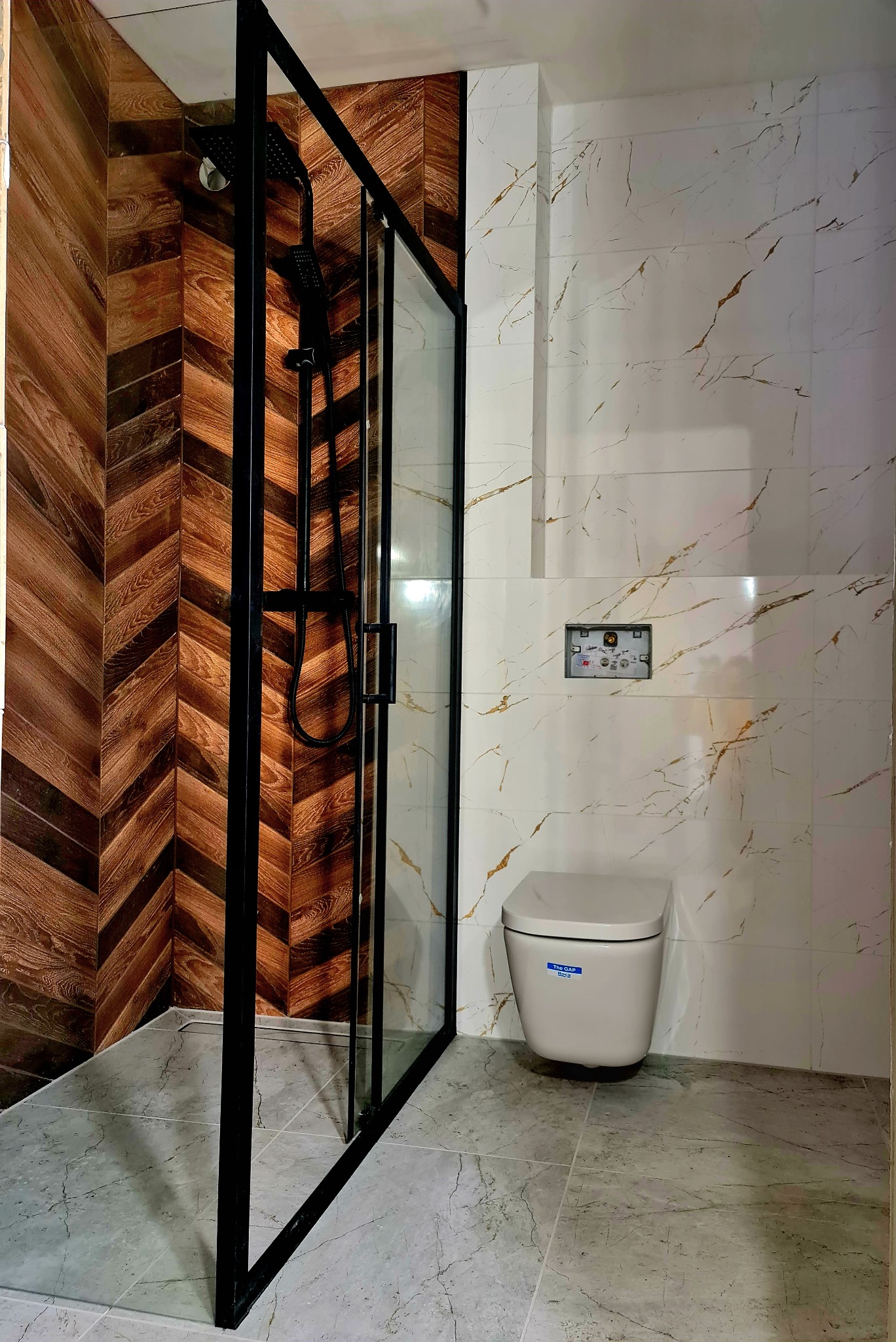 Nowoczesna łazienka z kabiną prysznicową z czarną ramą, drewnopodobną ścianą ułożoną w jodełkę, marmurowymi płytkami i podwieszaną toaletą.