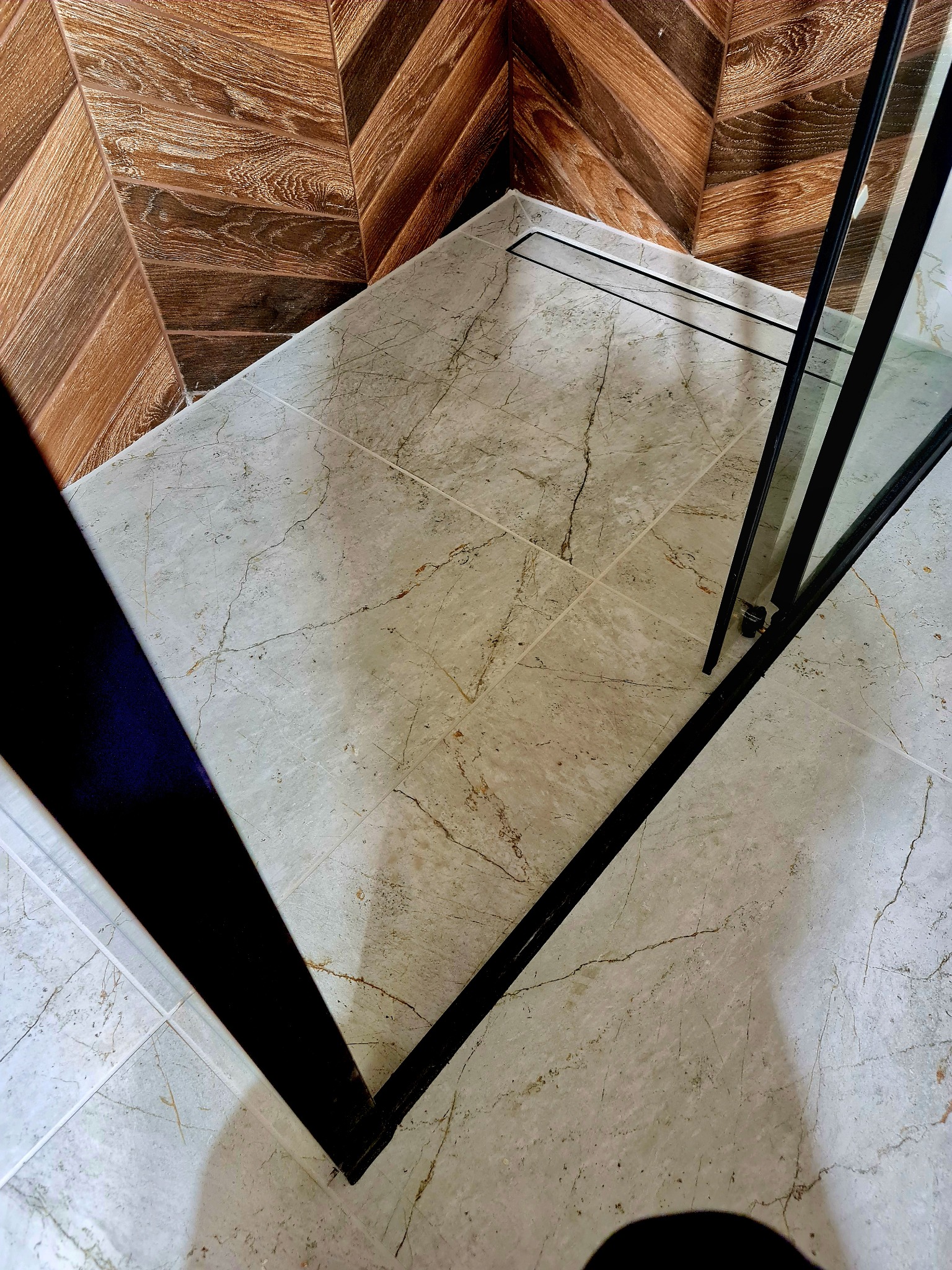 Wykończona łazienka z marmurową podłogą, odpływem liniowym i ścianą z drewnopodobnych płytek ułożonych w jodełkę, widok z góry na kabinę prysznicową z czarnymi profilami.